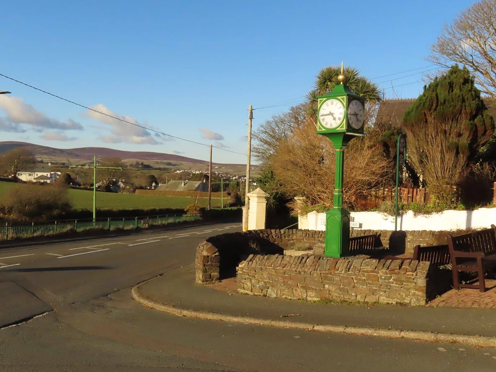 A green Victorian-style clock on a stone base at a road junction in Baldrine, with the hills of Lonan beyond.