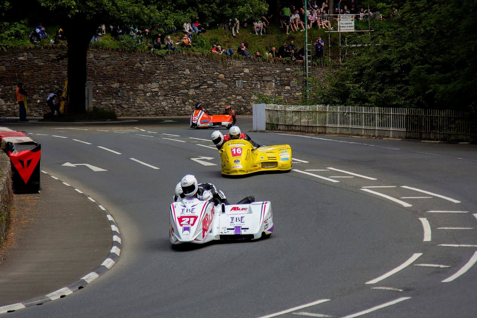 Two racing sidecar outfits leaning through the S-bend at Braddan Bridge on the TT course, with spectators on the bank behind.