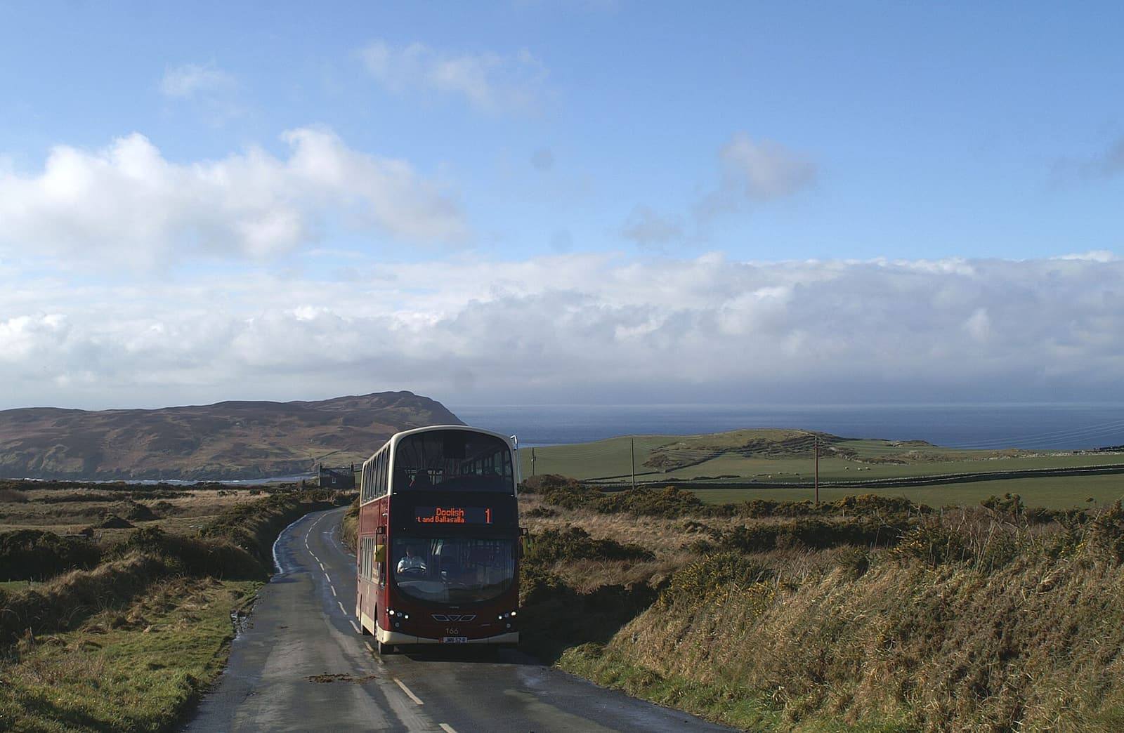 A double-decker bus on a narrow road through open, green and brown hills near Cregneash, with the sea behind.