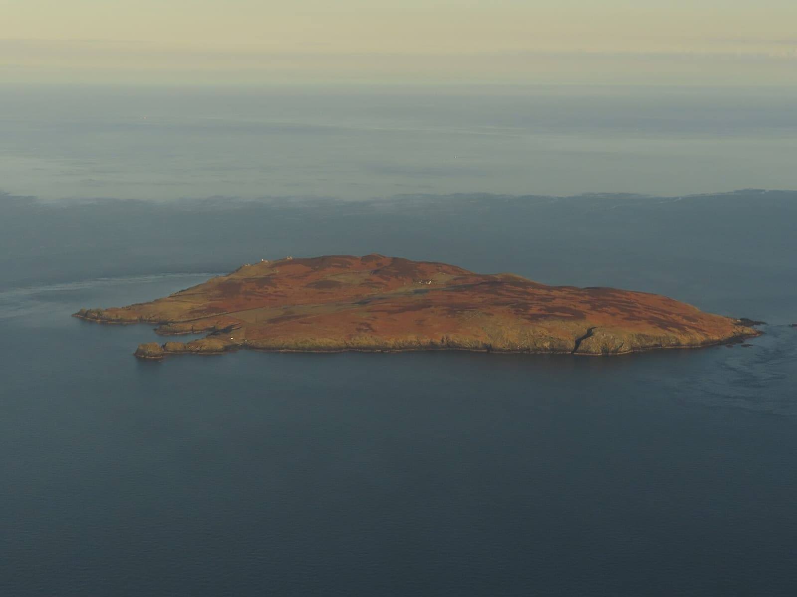 The Calf of Man seen from the window of a plane approaching Ronaldsway, surrounded by calm grey sea.