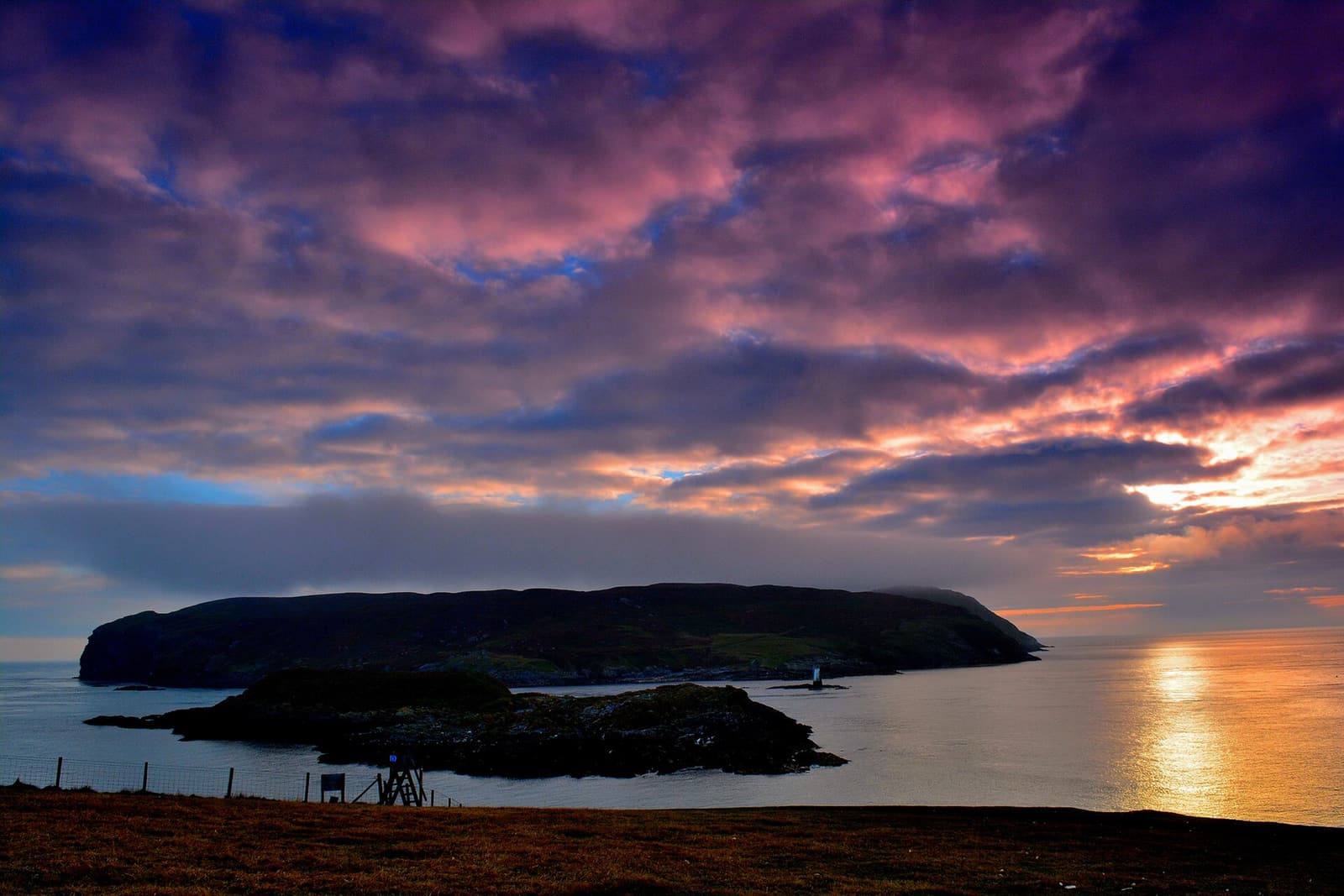 A dramatic purple and orange sunset sky over the dark outline of the Calf of Man and a calm sea.