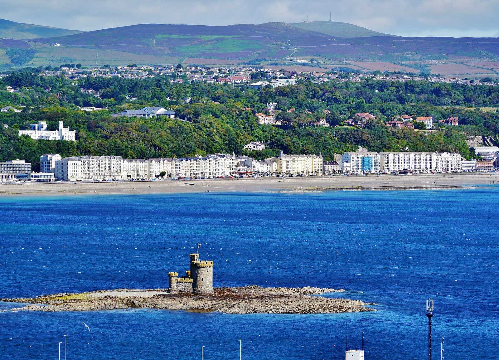 The hotels of Douglas promenade curve around the bay behind the Tower of Refuge on its rocky islet, with green hills beyond.