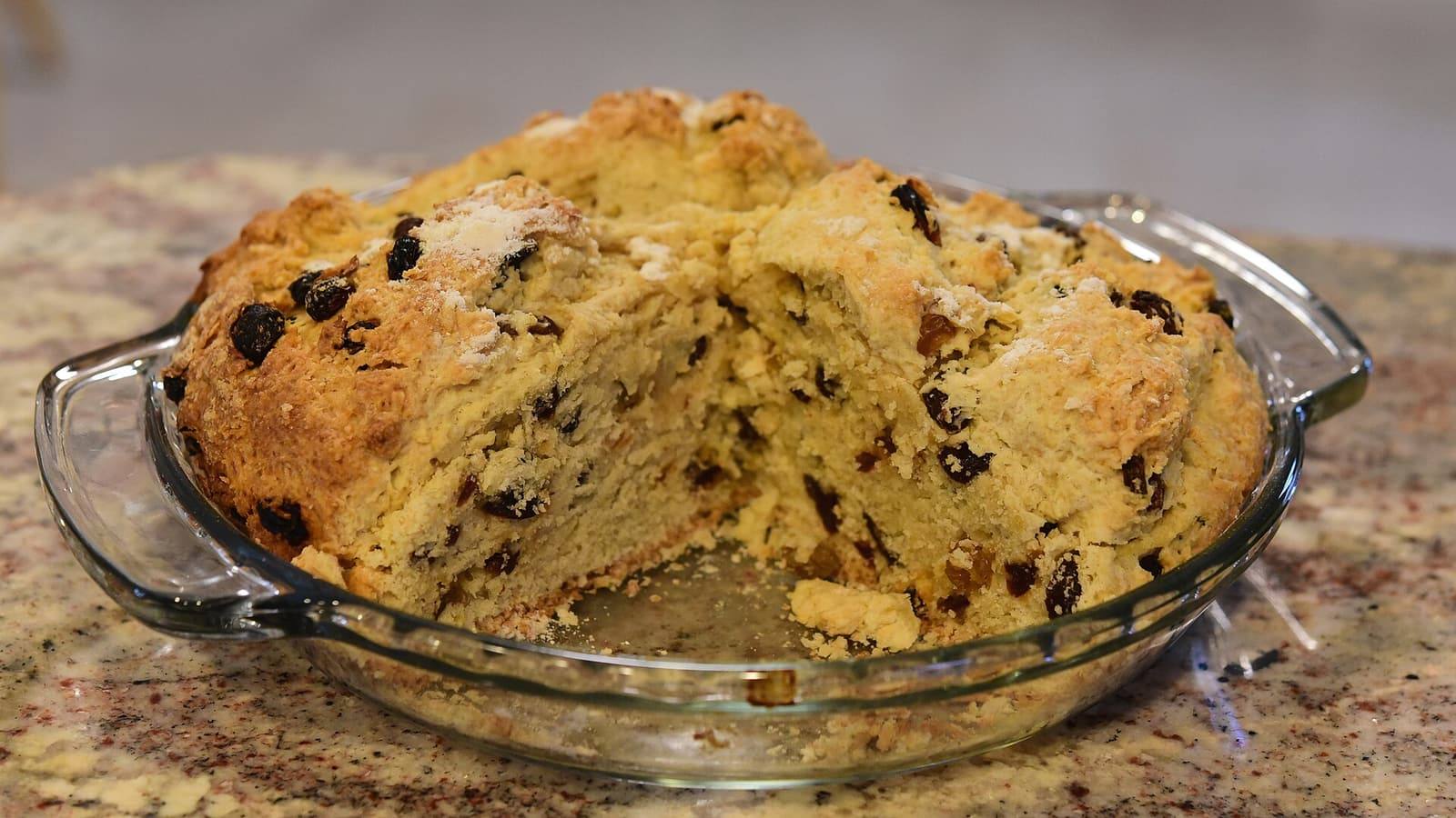 A round, golden soda bread dotted with raisins in a glass pie dish, with a wedge cut out.