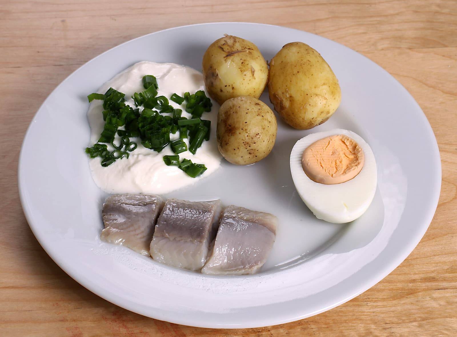 A white plate with three pieces of pickled herring, three small potatoes in their skins, sour cream topped with chives and half a boiled egg.