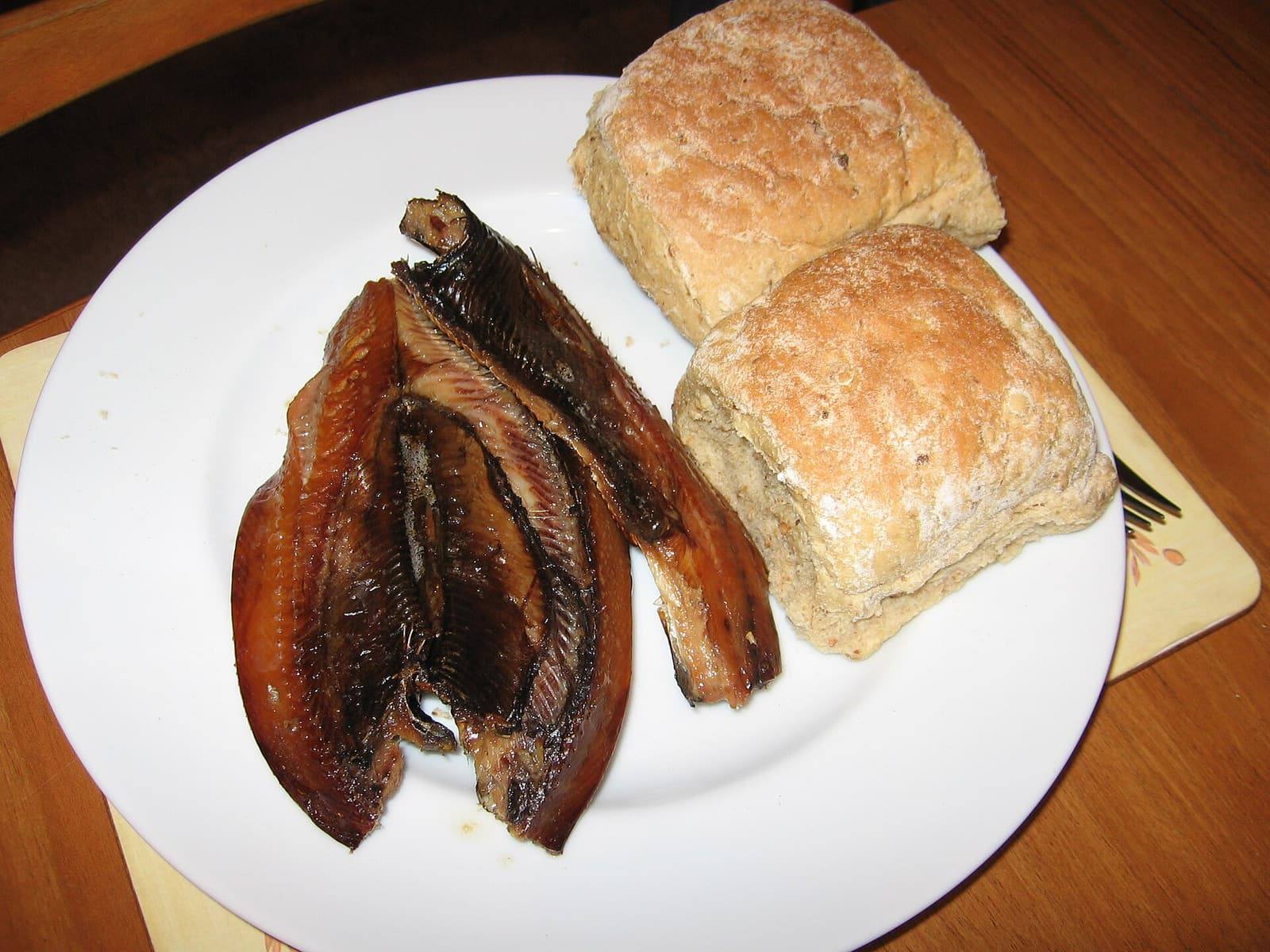 Two dark, glossy smoked kippers on a white plate beside two floury brown bread rolls.