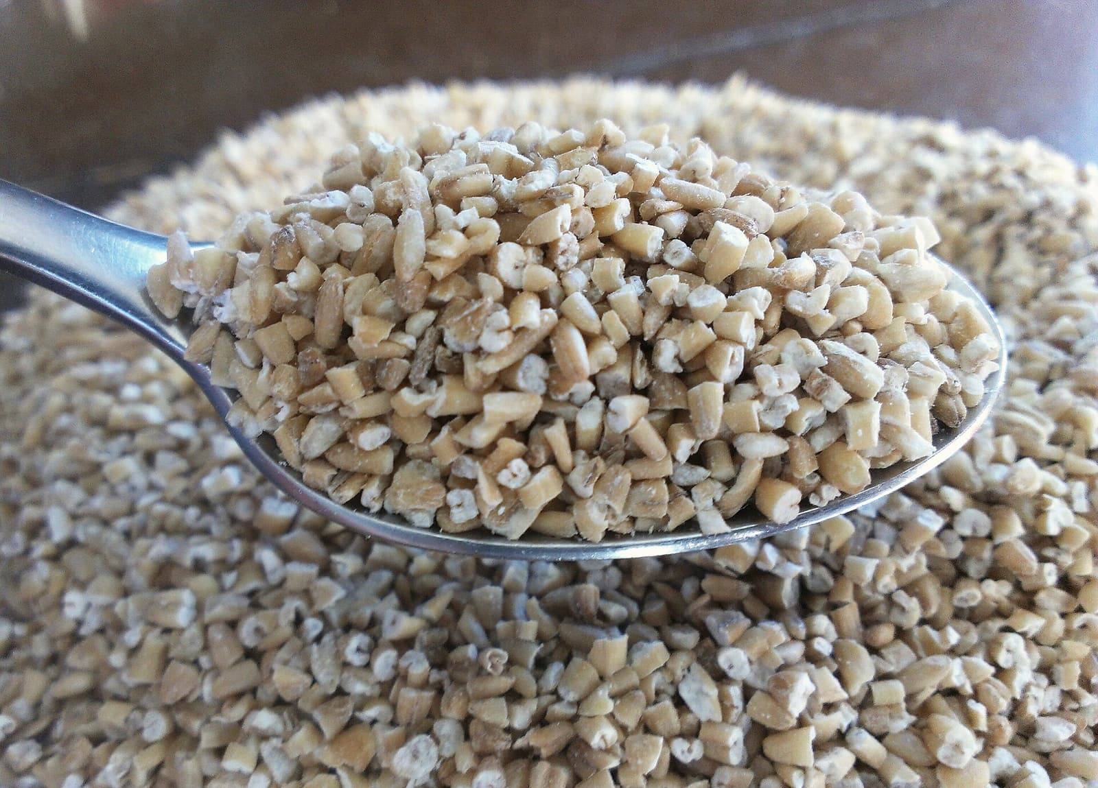 A close-up of a spoon heaped with dry, chopped oat grains over a bowl of more oats.