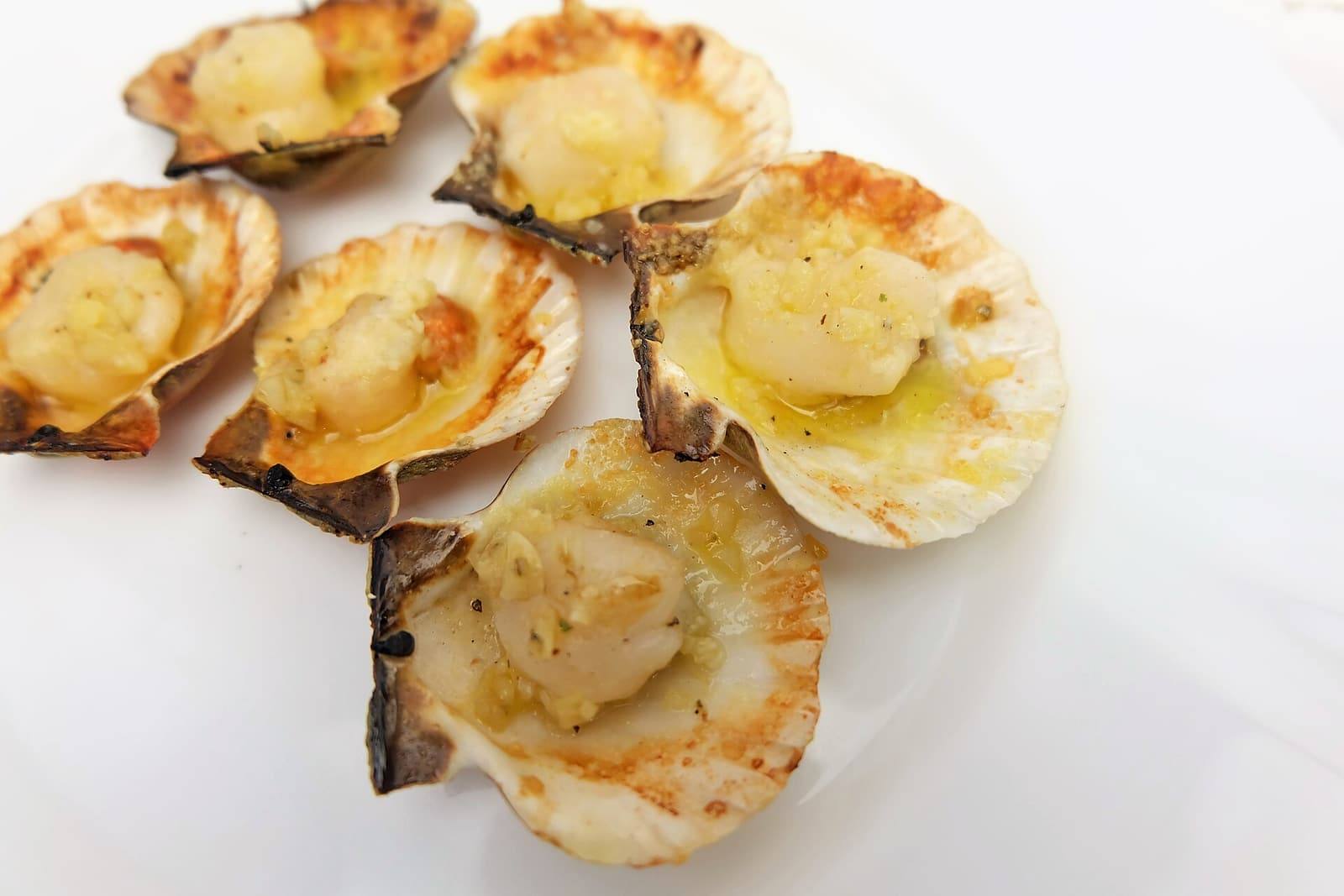 Six small scallops in their half shells, glistening with melted garlic butter, on a white plate.