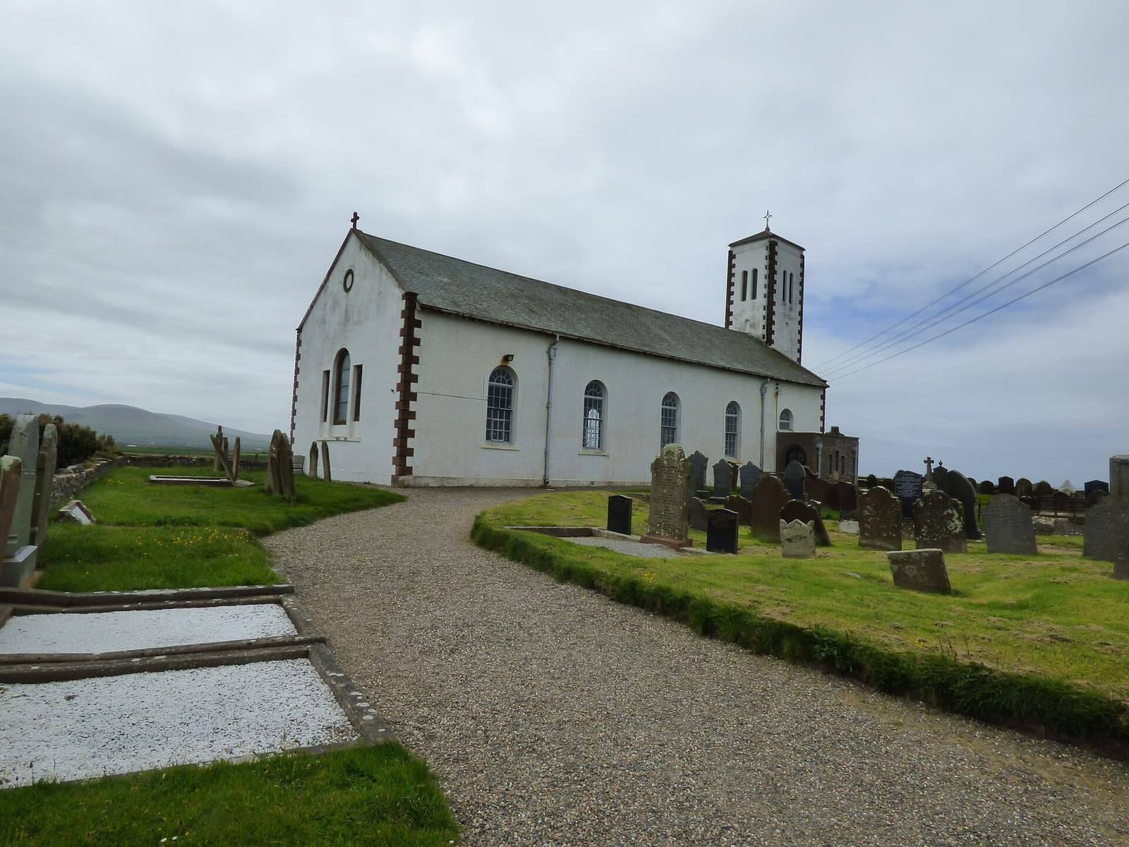 The white church at Jurby West with its square tower, reached by a gravel path between gravestones and grass.