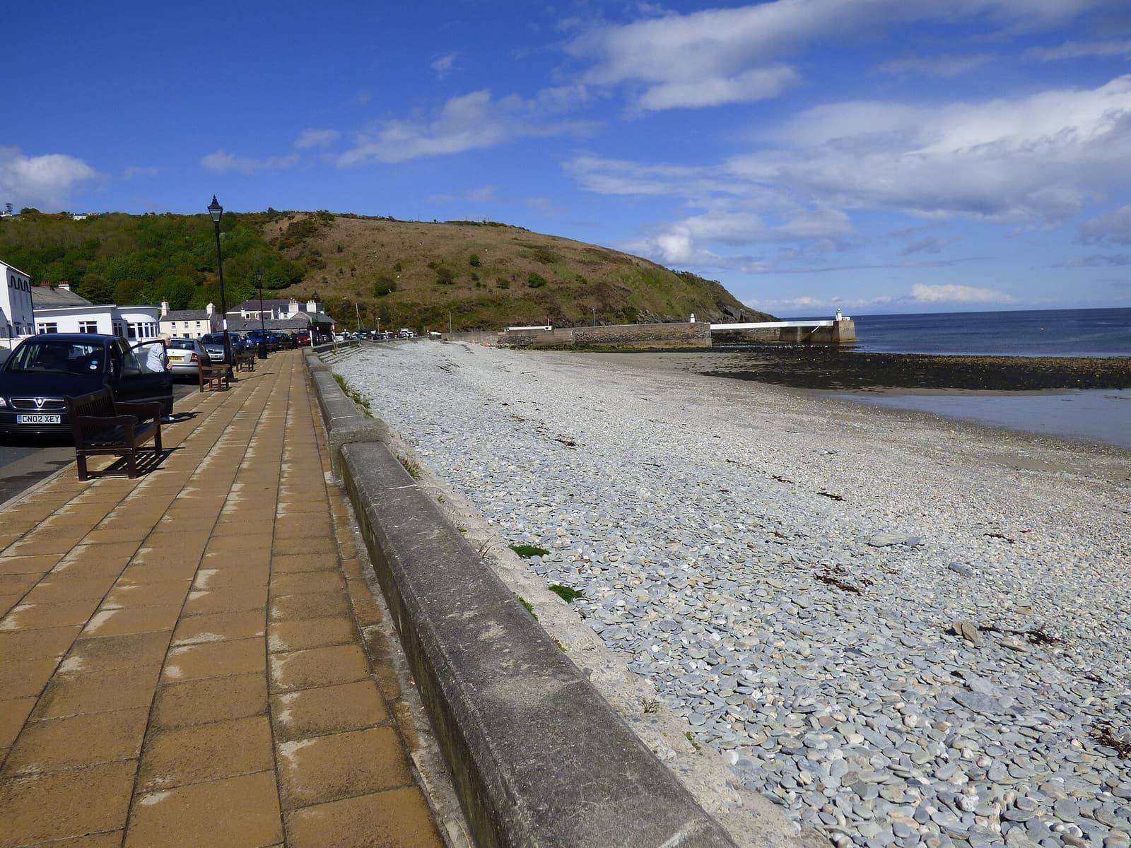 The promenade and shingle beach at Old Laxey running towards the harbour entrance and the headland on a sunny day.