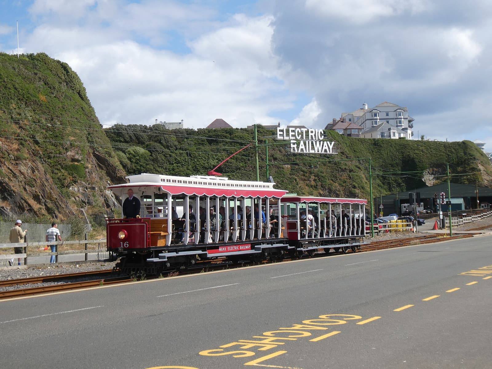 A red open-sided Manx Electric Railway tram near Derby Castle at the north end of Douglas promenade, below the cliffs.