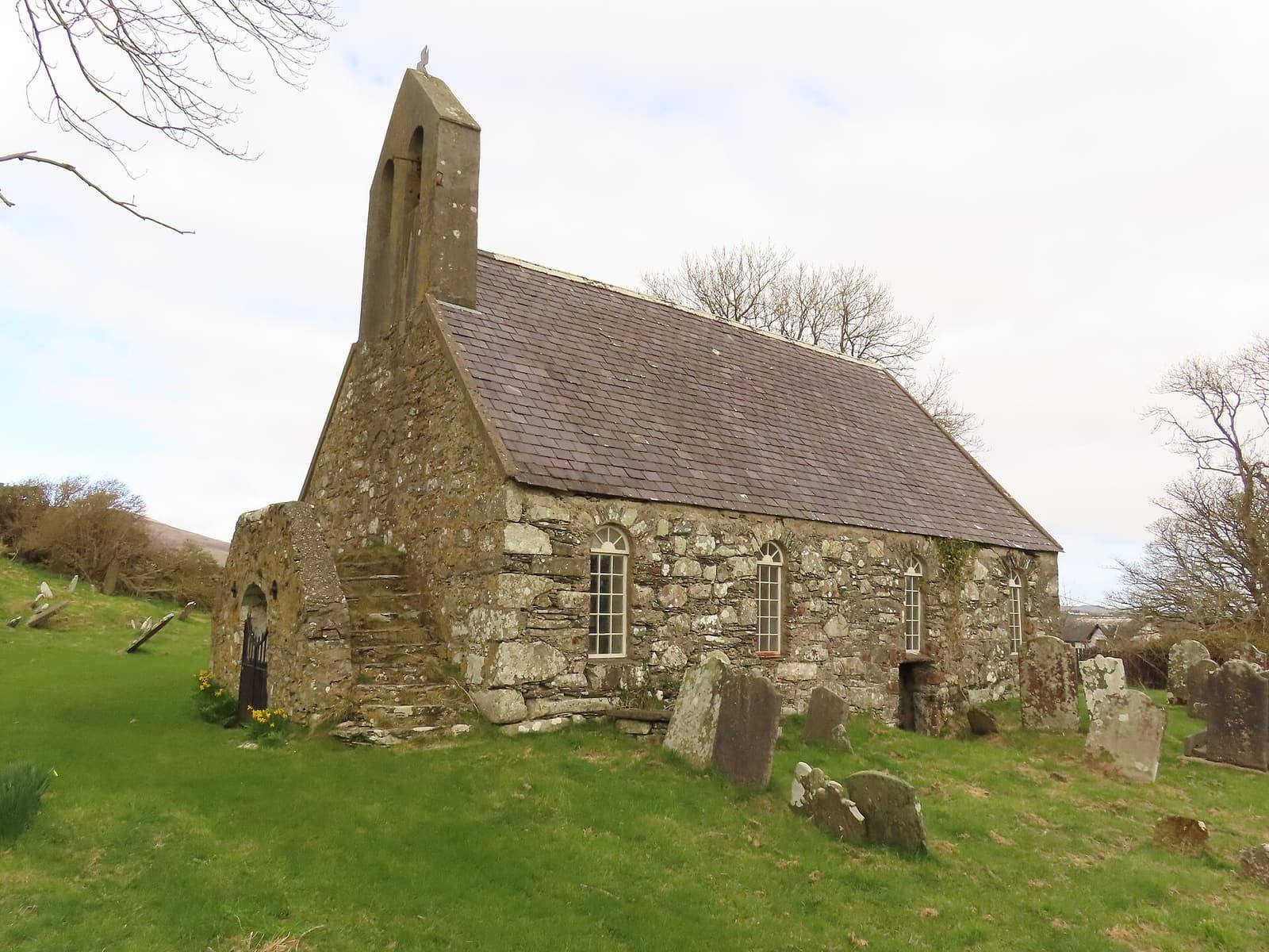 The small stone Marown Old Church with its bell-cote, on a grassy rise among old gravestones under a bright sky.