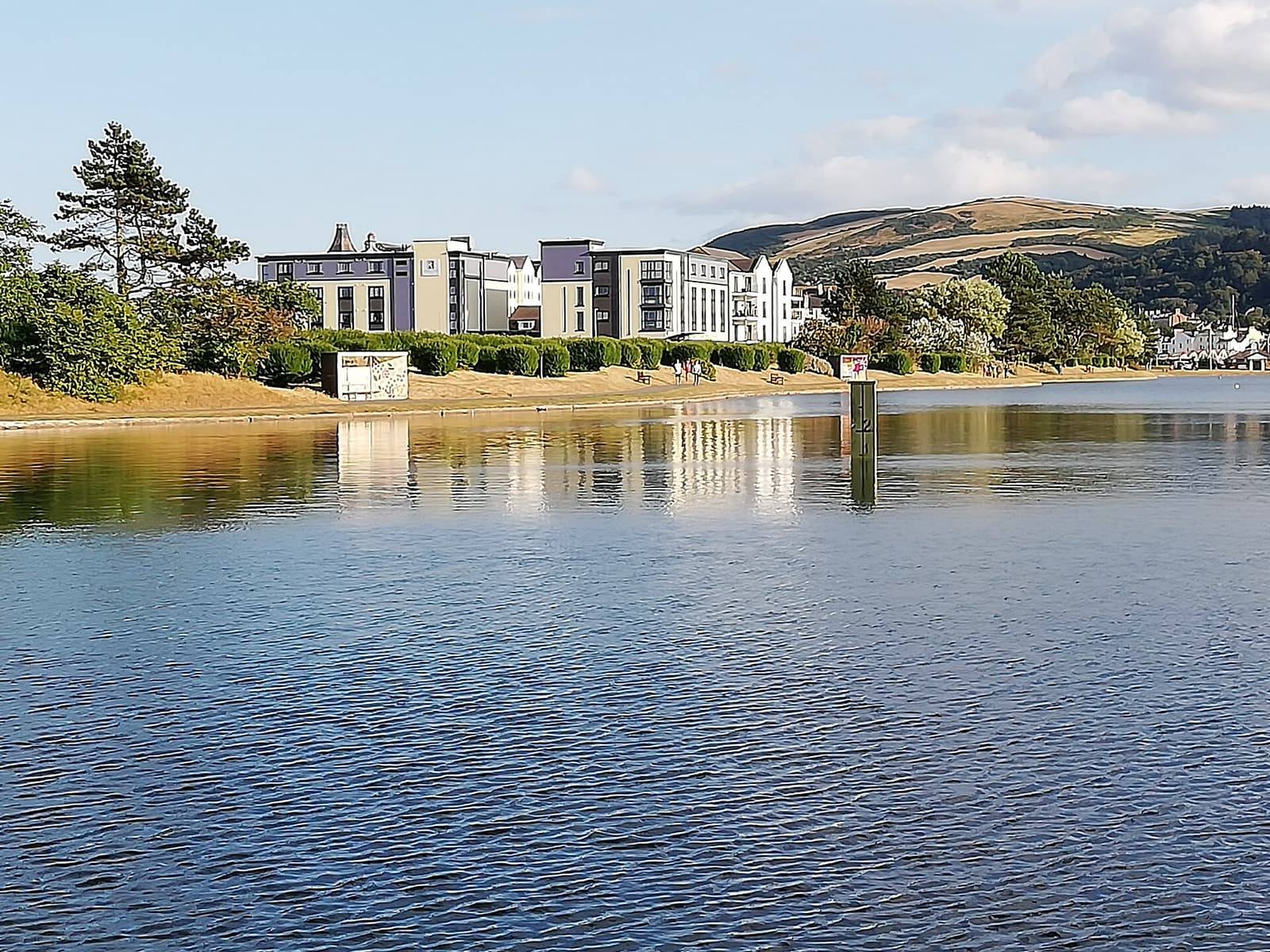 The calm boating lake at Mooragh Park in Ramsey with trees on its far bank and hills behind.