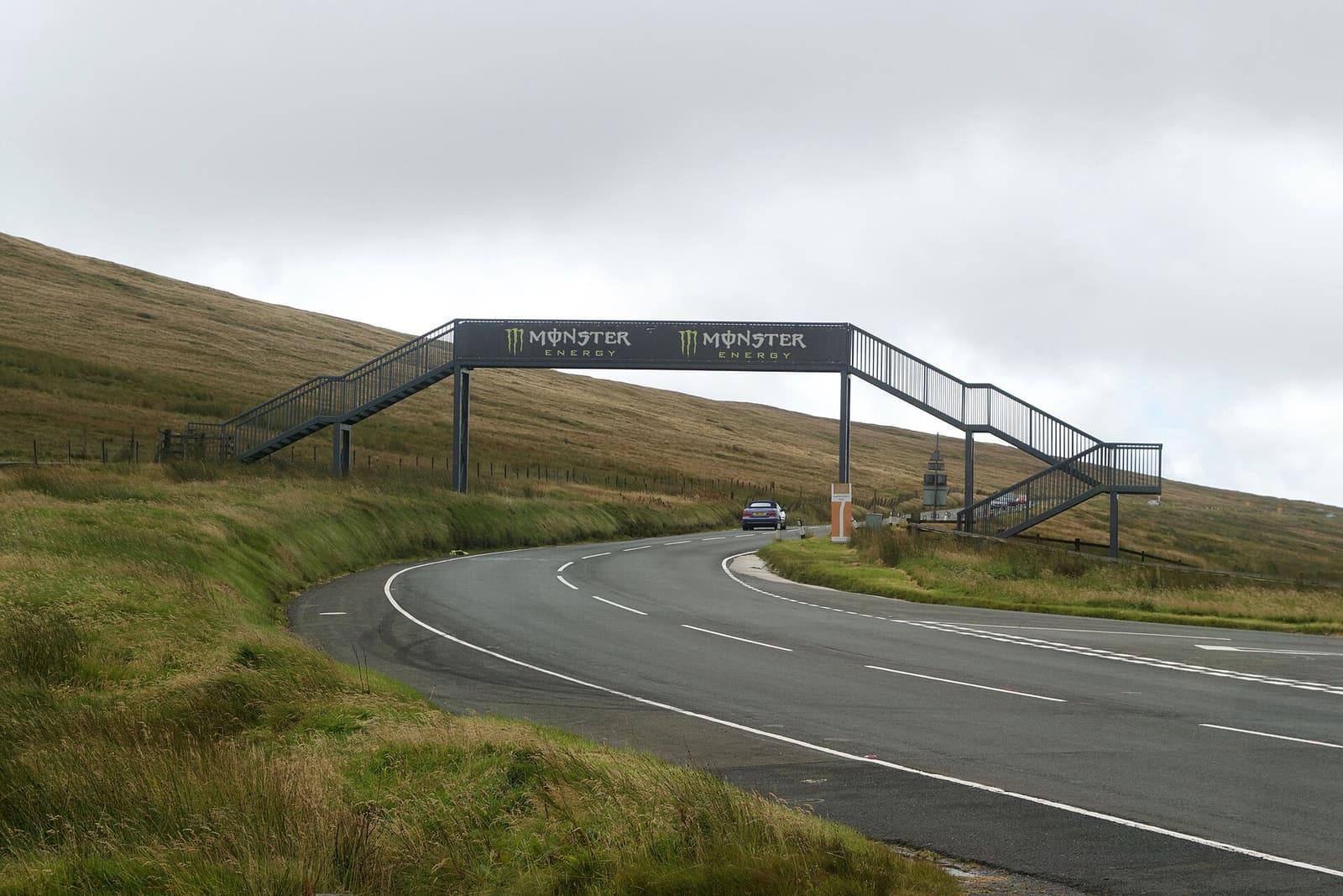 The Mountain Road curving across open moorland under a footbridge built for the TT, beneath a grey sky.
