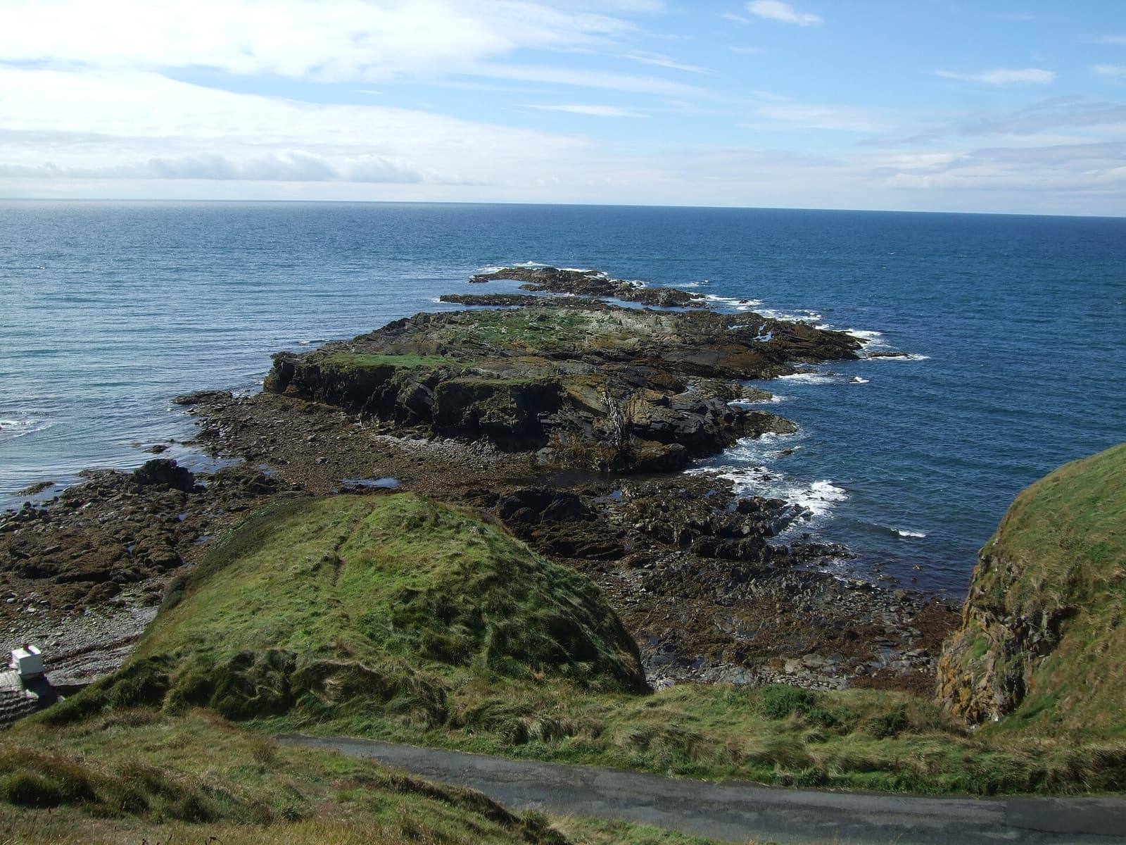 The rocky tail of Niarbyl reaching out into a blue sea, with green slopes and the coast stretching north.