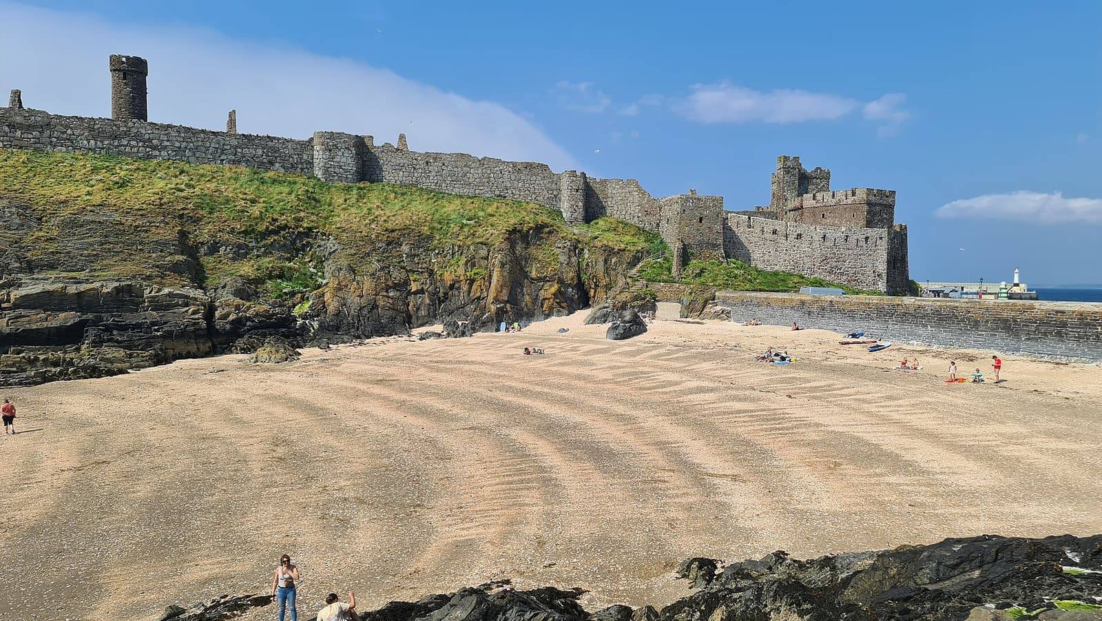 The walls of Peel Castle on St Patrick's Isle above the golden sand of Fenella Beach under a blue sky.