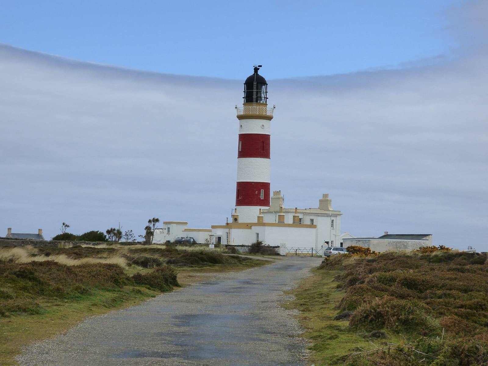 A narrow road through rough grass leading to the tall red-and-white Point of Ayre lighthouse and its white buildings.