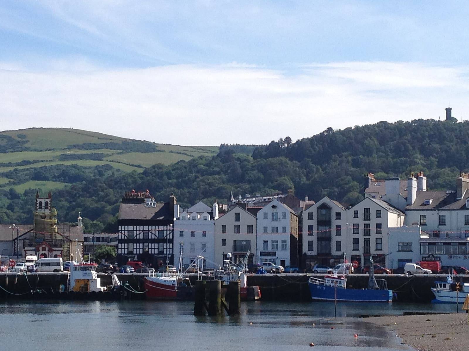 Fishing boats in Ramsey harbour in front of the quayside buildings, with wooded hills and the Albert Tower behind.
