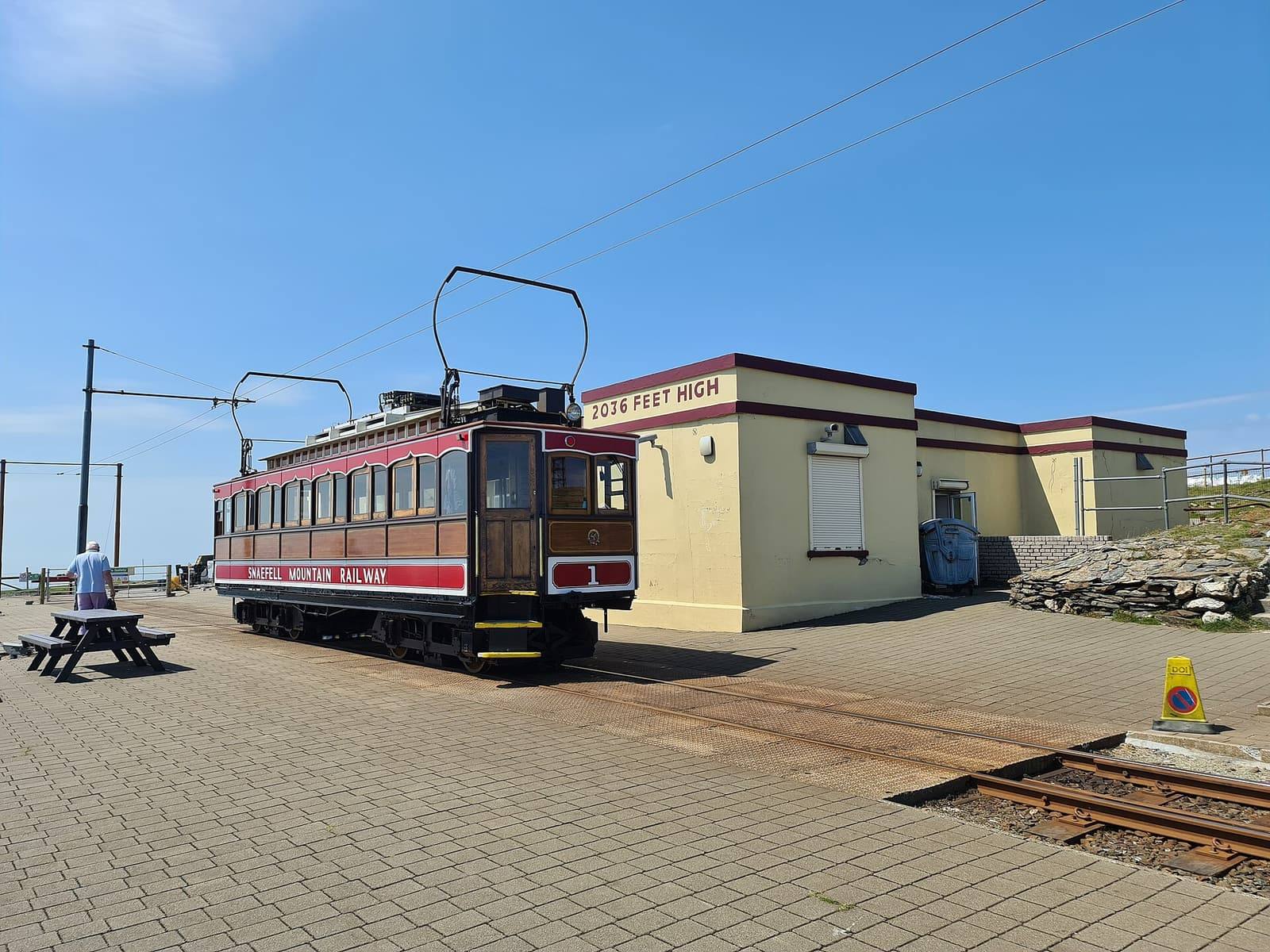 A red and cream Snaefell Mountain Railway car beside the summit station building under a clear blue sky.
