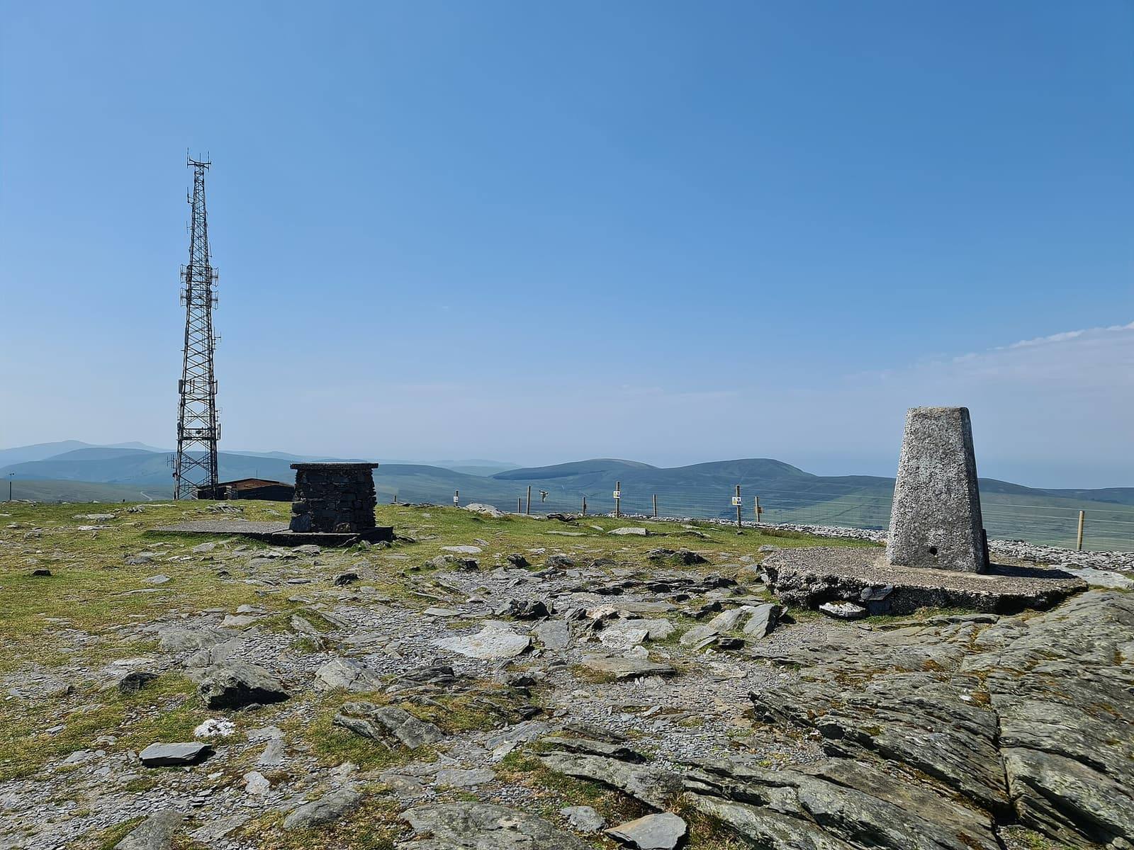 The white trig pillar on the rocky summit of Snaefell, with views across the island's hills under a blue sky.