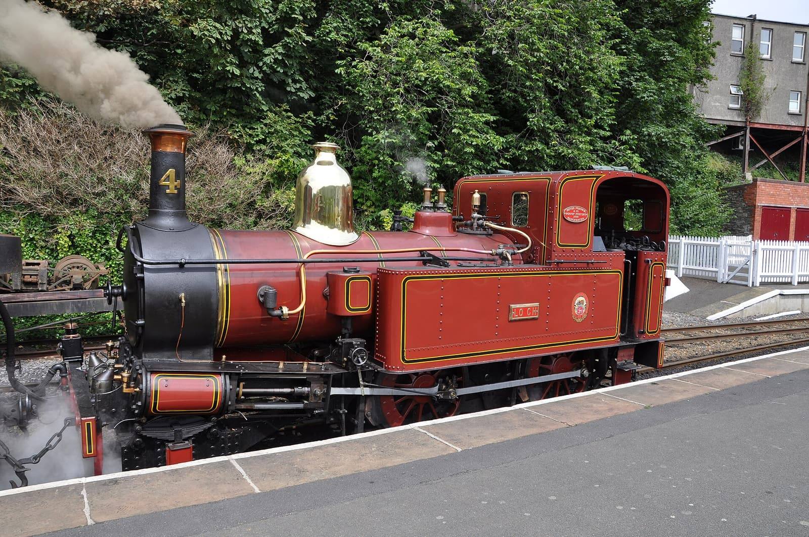 A red Isle of Man Railway steam engine with a polished brass dome letting off steam at Douglas station.