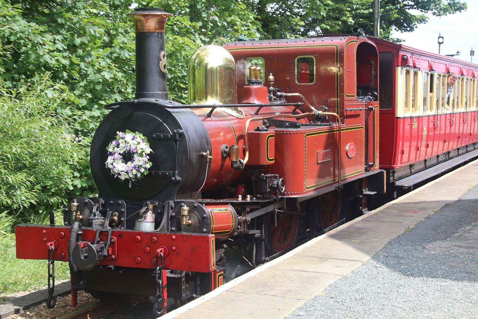 A red Isle of Man Railway steam engine pulling red carriages into Port St Mary station between green trees.