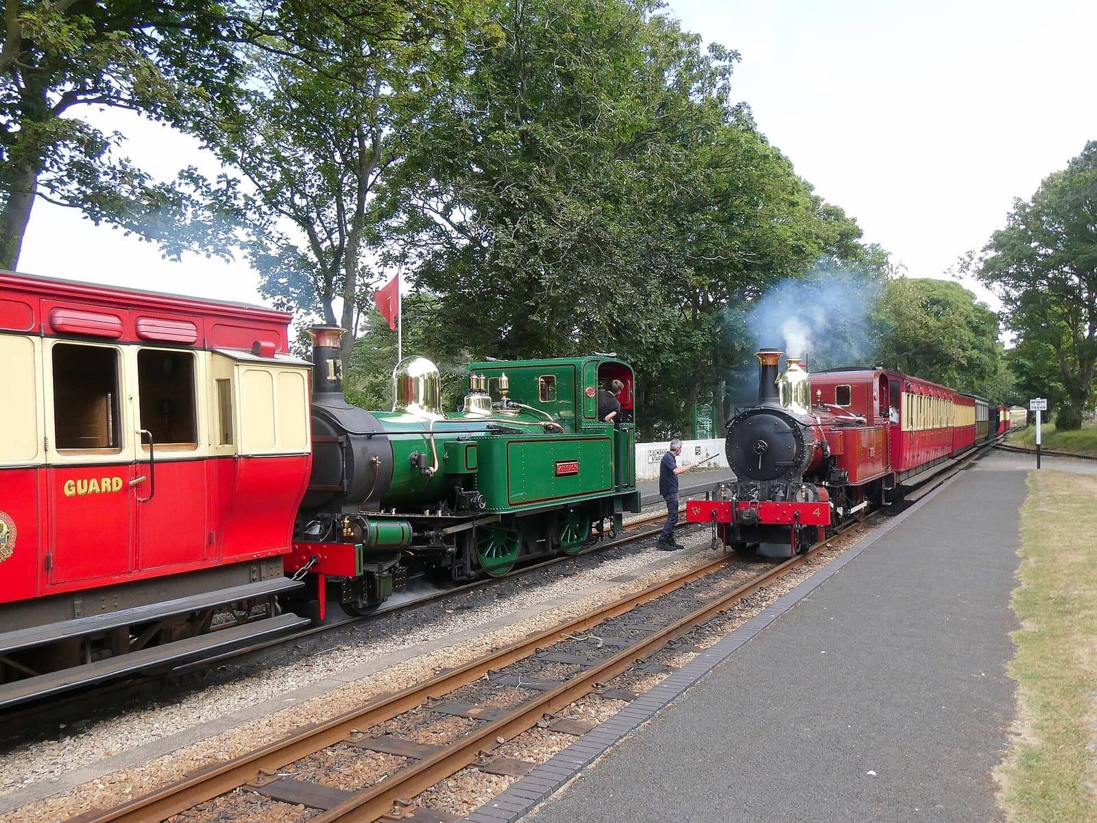 A green and a red steam engine with red carriages passing each other at Castletown station in summer.