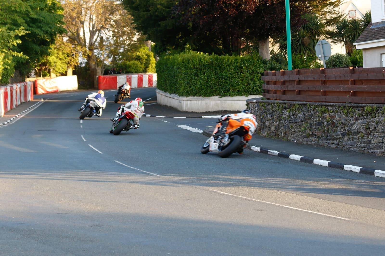 Three motorcycle racers leaning through Cruickshank's Corner on May Hill in Ramsey, with straw-bale and fence protection at the roadside.