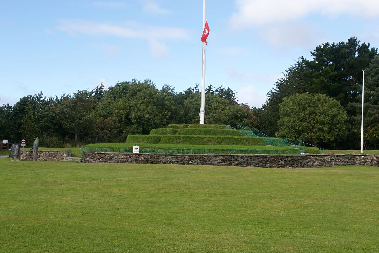 The stepped, grass-covered mound of Tynwald Hill at St John's with a flagpole at the top and trees behind.