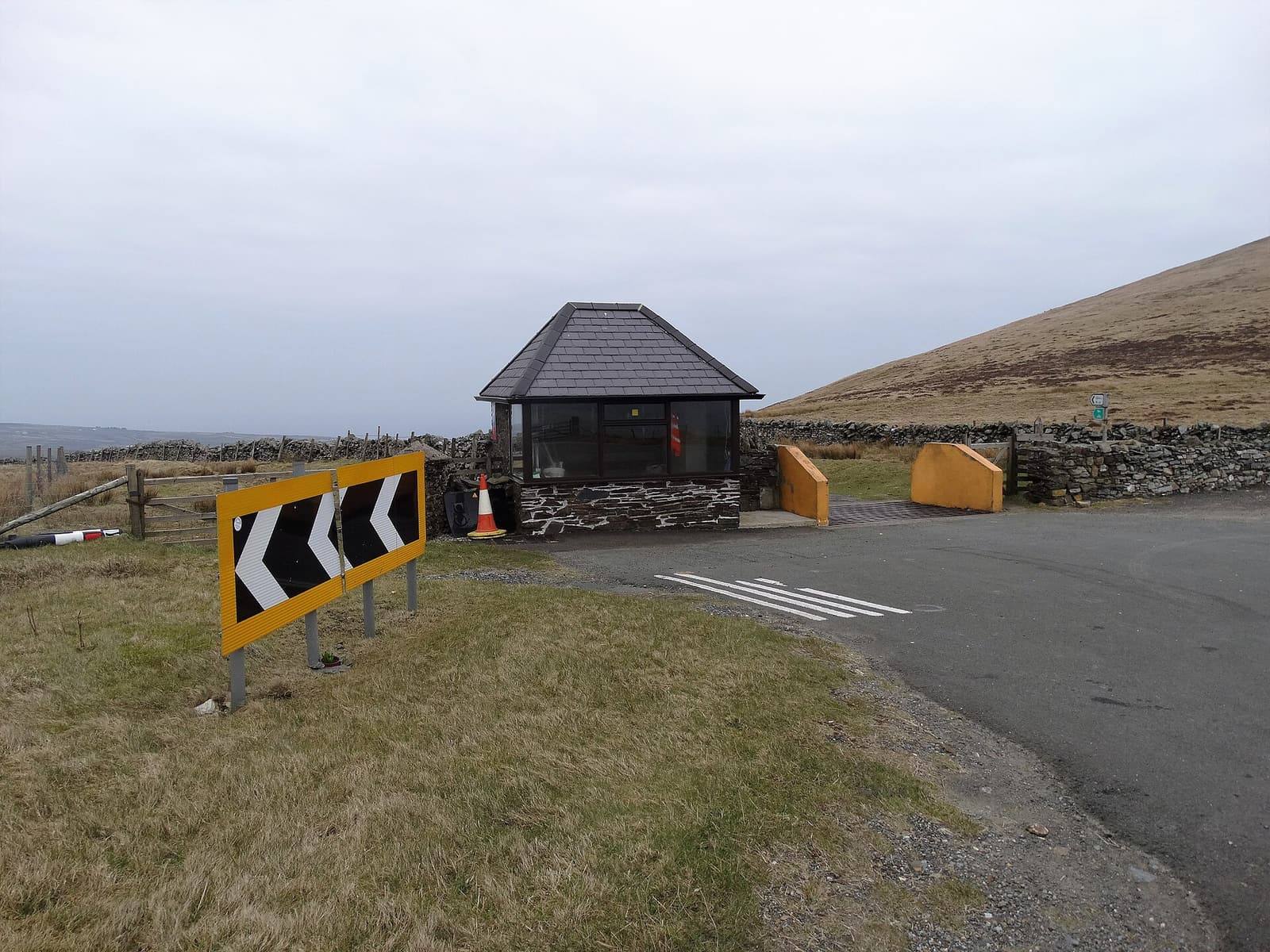 The Mountain Road bending at Windy Corner, with a chevron sign, a small stone shelter and open hillside behind.